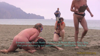 Two naked guys are “Wrestling clothing-free at a beach festival”. Also, topless photographer’s boobs are visible in the background